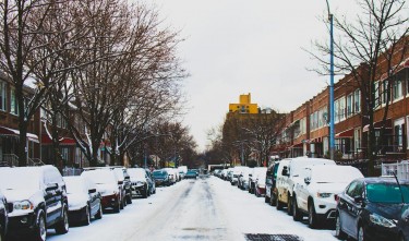 SNOW COVERED ROAD NAD INLINE PARKED VEHICLES BETWEEN 2 STOREY BUILDINGS UNDER WHITE SKY